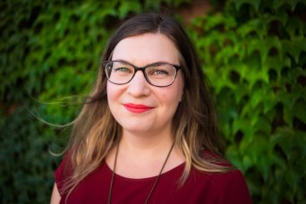 Photo of a white woman with long brown hair, wearing glasses and a maroon shirt, standing against a brick wall with green vines